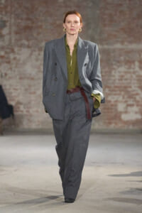 Model on a runway wearing a loose gray double-breasted suit with an olive green shirt and brown belt.