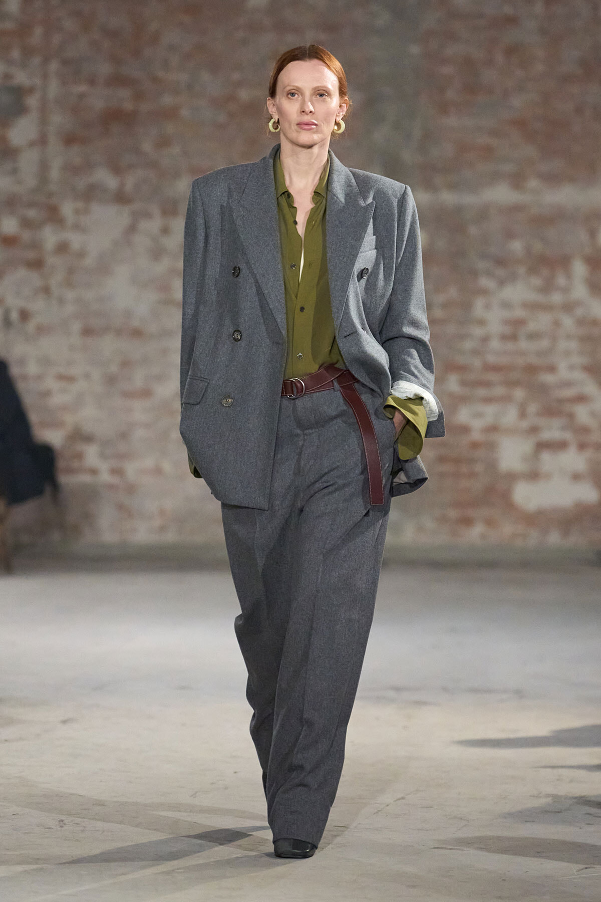 Model on a runway wearing a loose gray double-breasted suit with an olive green shirt and brown belt.