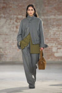 Model walks a runway in a gray pinstripe suit with an oversized wrap jacket and olive-green inner panels, carrying a brown suede bag.
