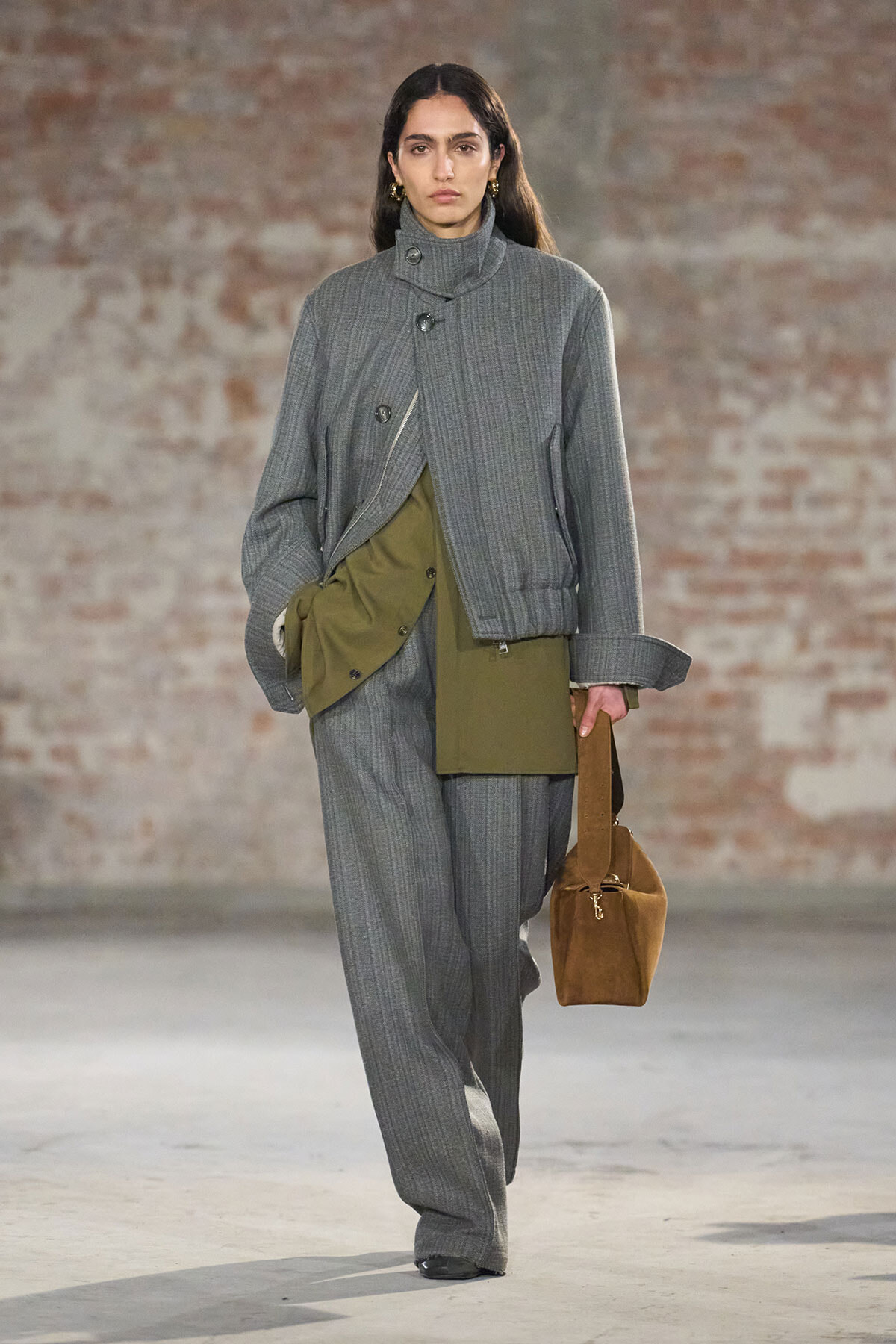 Model walks a runway in a gray pinstripe suit with an oversized wrap jacket and olive-green inner panels, carrying a brown suede bag.