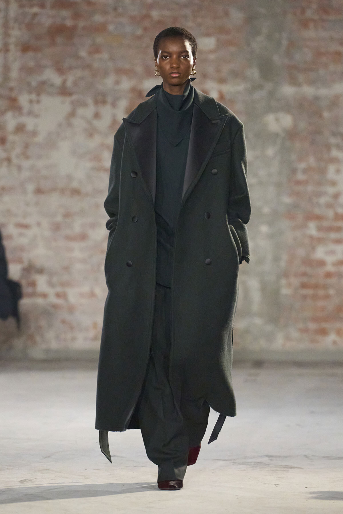 Model on a runway wearing a long dark green double‑breasted coat over a black outfit, with a brick wall backdrop.