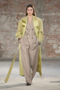 Model walks the runway in a beige double-breasted suit with wide-leg trousers, paired with a long pale-green trench coat and hands in pockets.