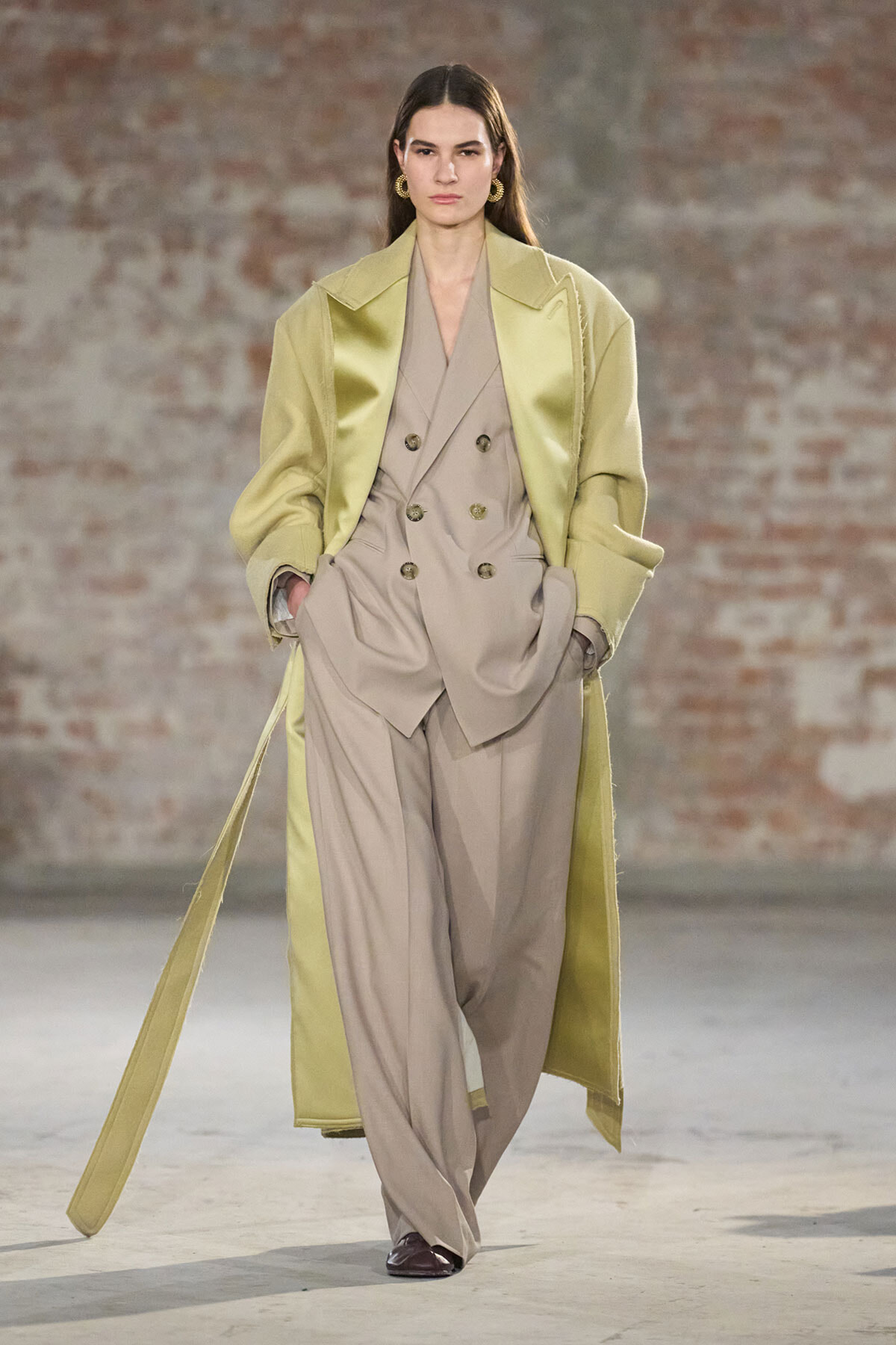 Model walks the runway in a beige double-breasted suit with wide-leg trousers, paired with a long pale-green trench coat and hands in pockets.