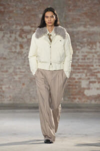 Female model walks a runway in a cream puffer jacket with a gray fur collar and loose beige wide-leg pants, brick wall background.