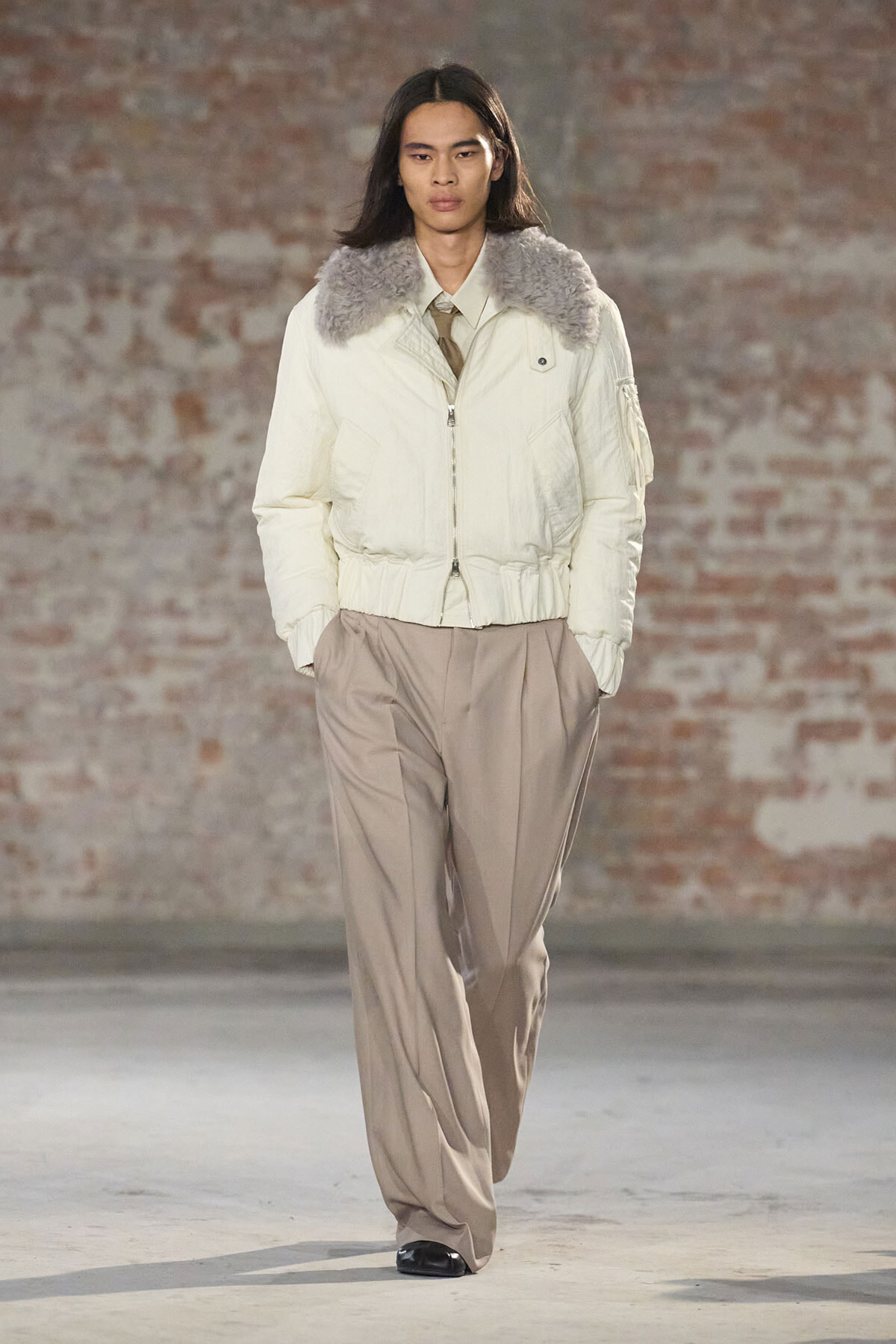 Female model walks a runway in a cream puffer jacket with a gray fur collar and loose beige wide-leg pants, brick wall background.