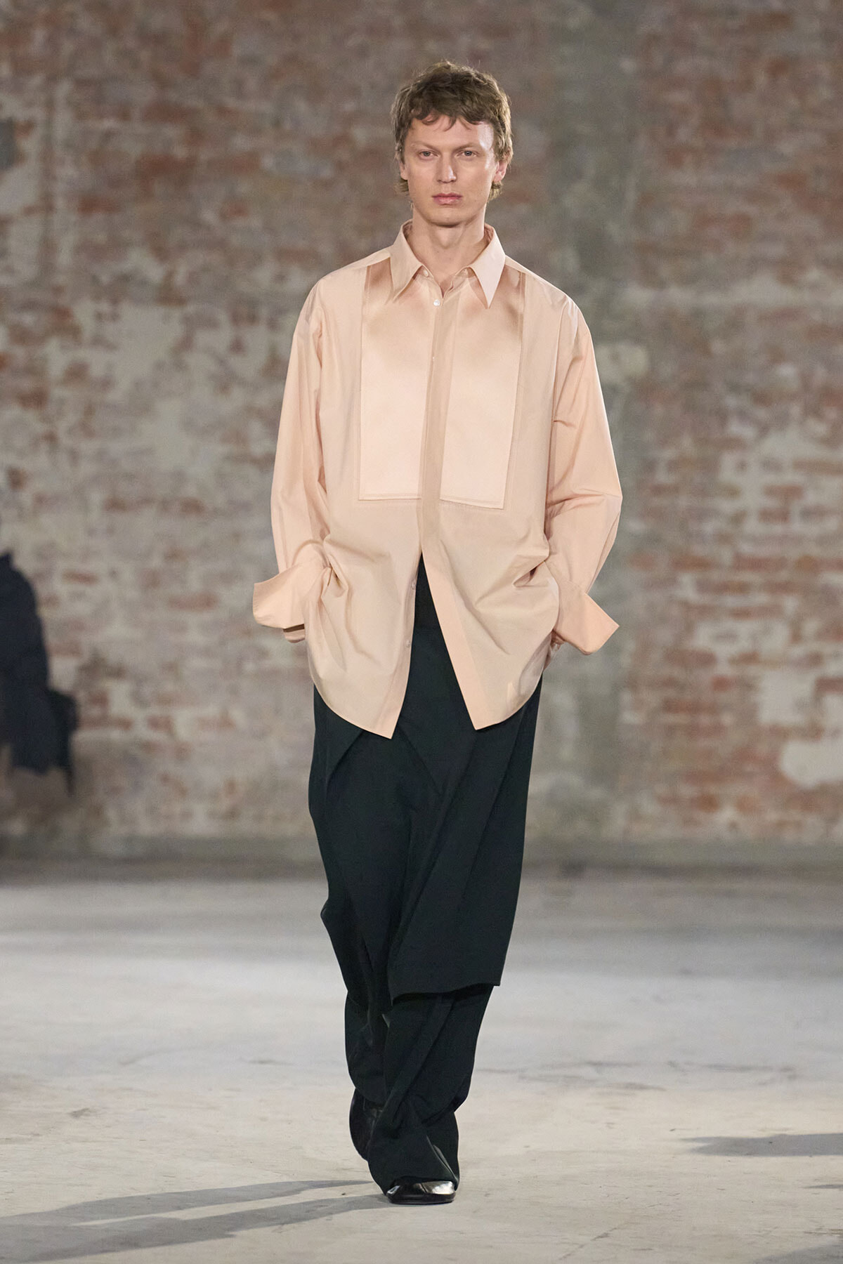 Model walks a runway in an oversized peach shirt and wide black trousers with a brick wall backdrop.