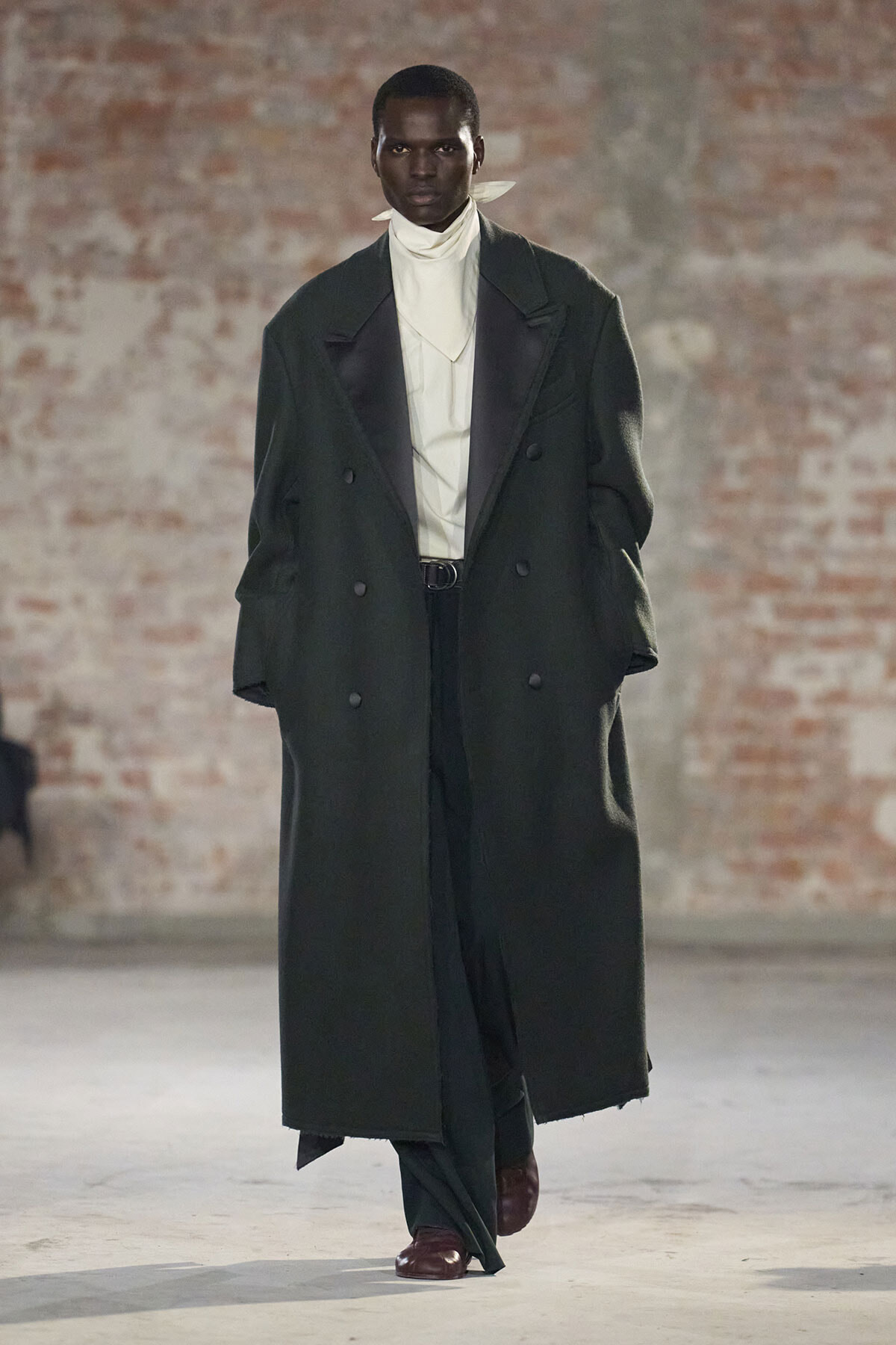 Male model walks a runway in a long dark green double-breasted coat, white turtleneck, black pants, and burgundy shoes.
