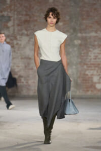 Model walking the runway in a white sleeveless top and a flowing gray wrap skirt, with black knee-high boots and a light blue handbag.