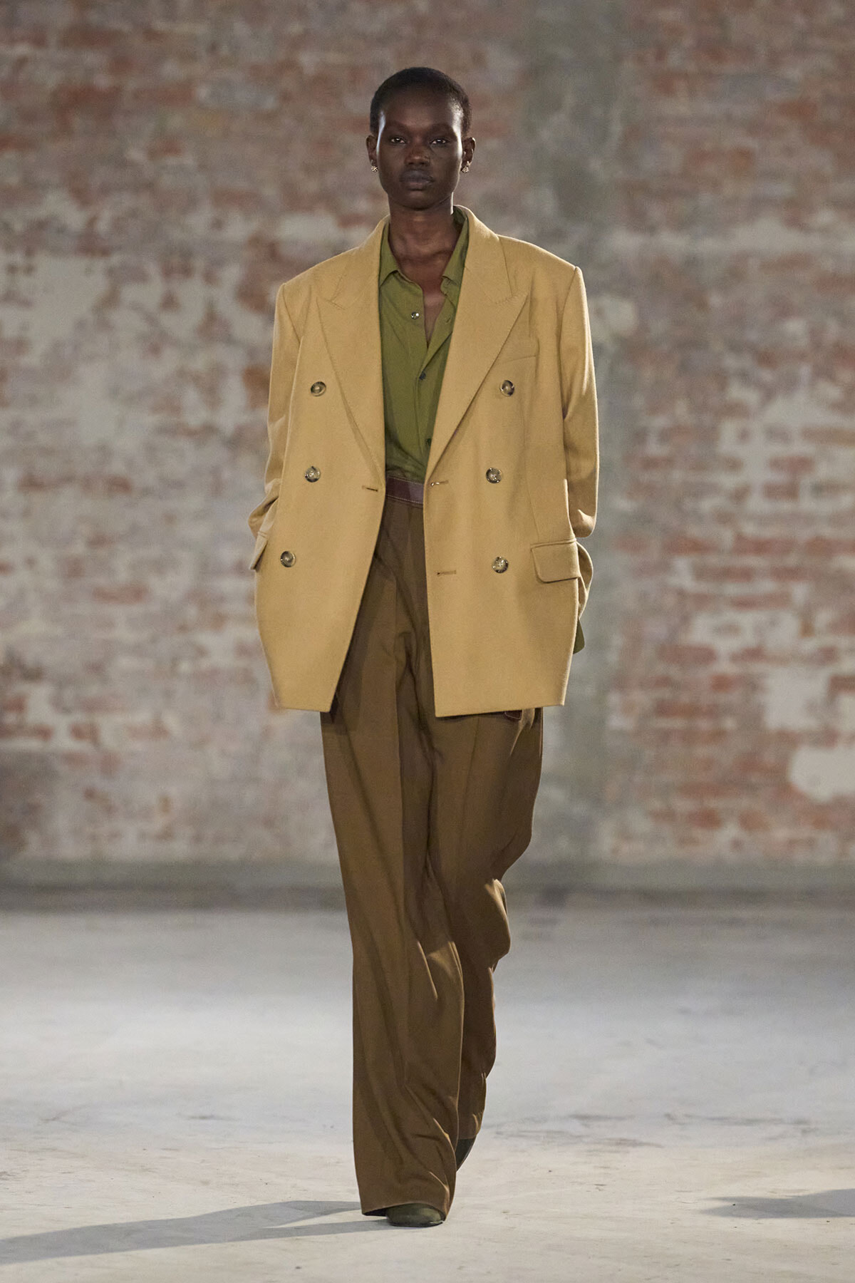 Model on a runway wearing a beige double-breasted blazer over an olive green shirt and brown wide-leg pants in an industrial brick setting, hands in pockets.