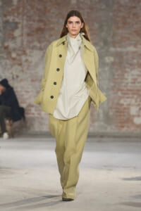Model on runway in oversized beige coat, cream blouse, and wide beige pants with a brick backdrop.