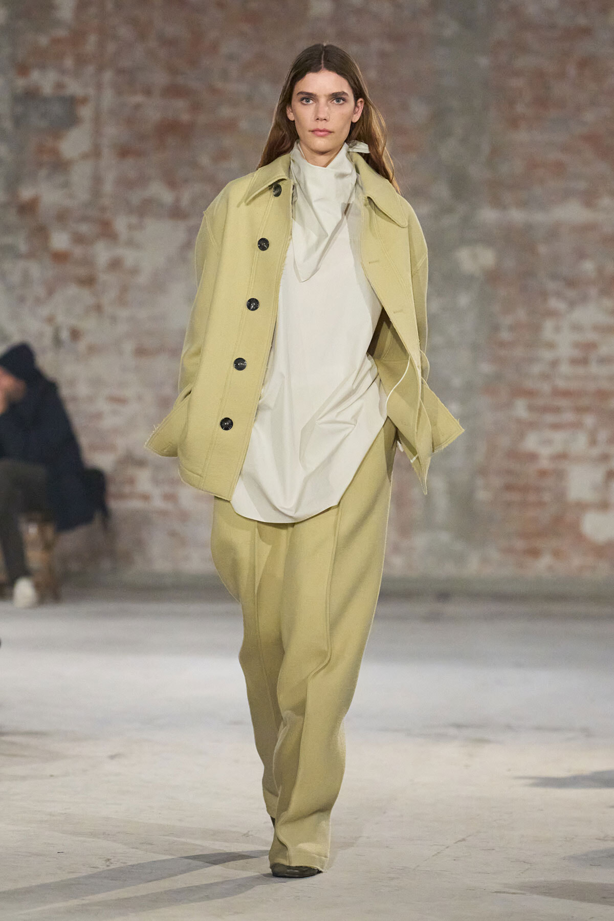 Model on runway in oversized beige coat, cream blouse, and wide beige pants with a brick backdrop.