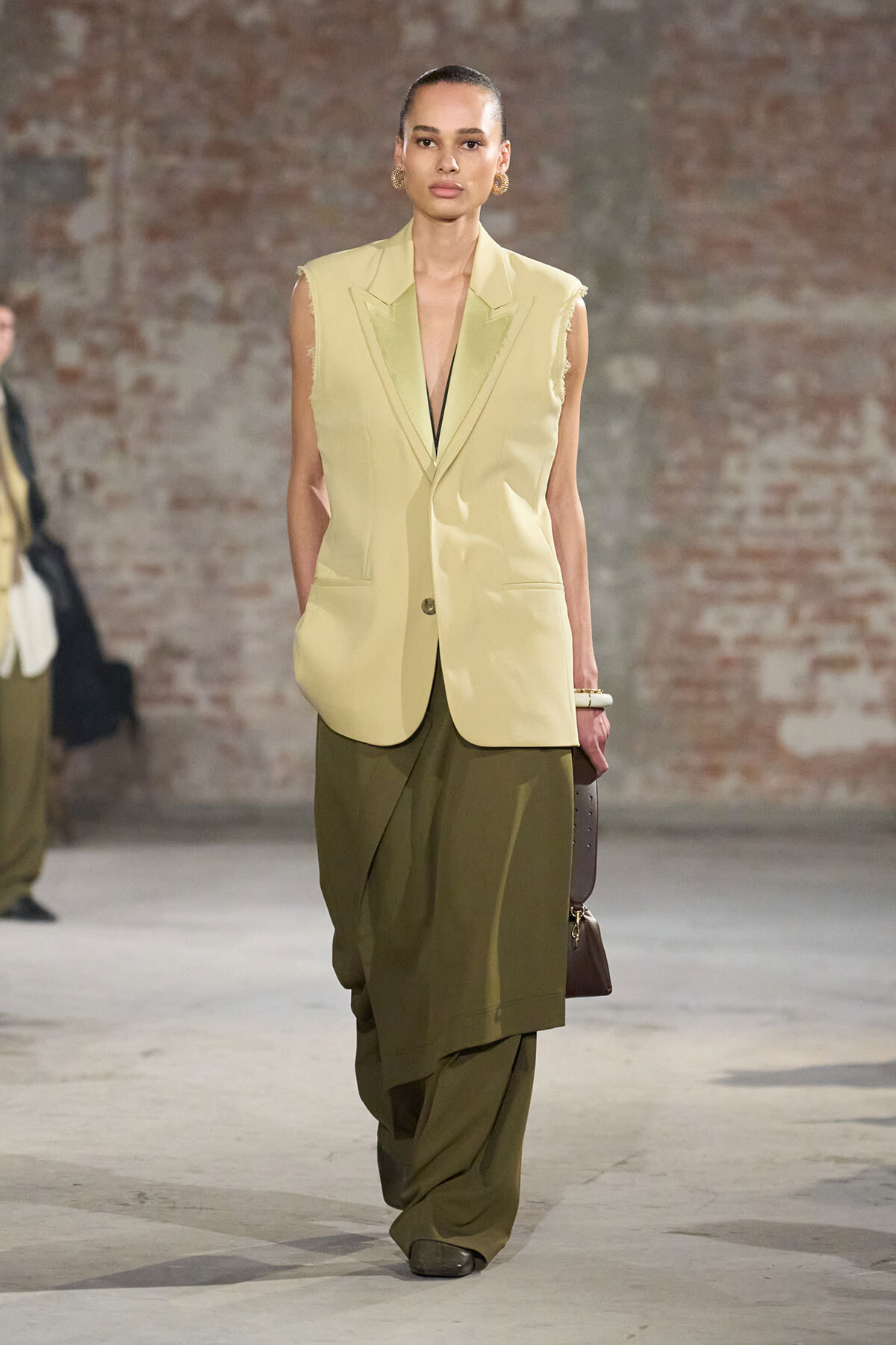 Model walking a runway in a pale yellow sleeveless blazer over olive draped trousers, brick-wall backdrop, minimalist look.