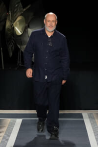 Man with a gray beard walking down a fashion runway in a dark navy shirt and pants, glasses hanging from chest.