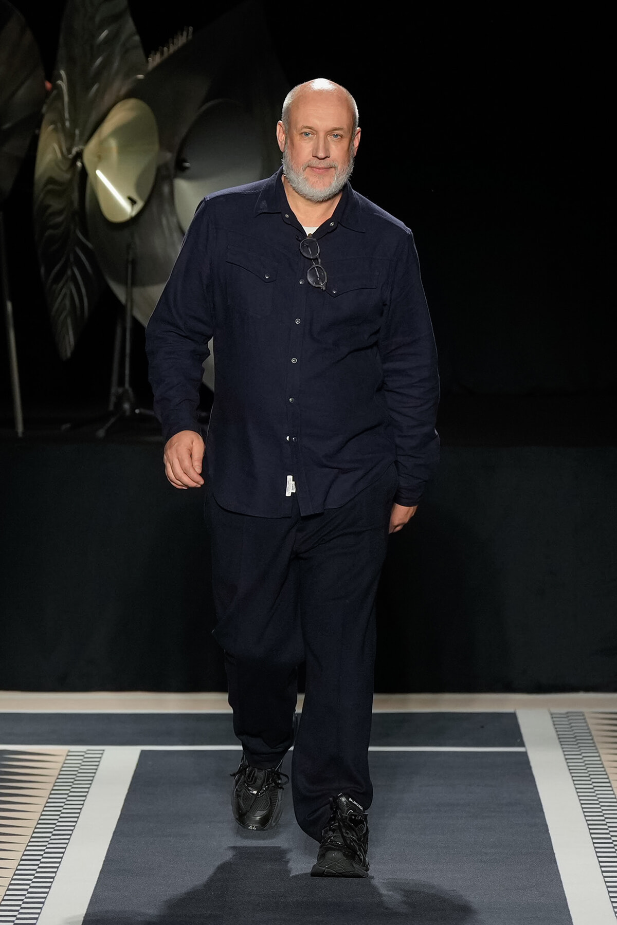 Man with a gray beard walking down a fashion runway in a dark navy shirt and pants, glasses hanging from chest.
