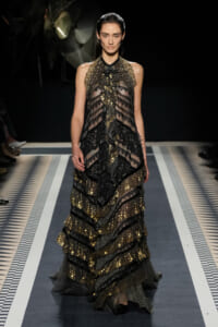 Model walking a fashion runway in a floor-length black lace gown with gold embellishments and tiered ruffles.