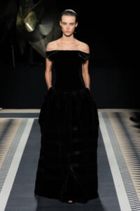 Model on a runway wearing a black off‑the‑shoulder velvet gown with a fitted bodice and full floor‑length skirt.