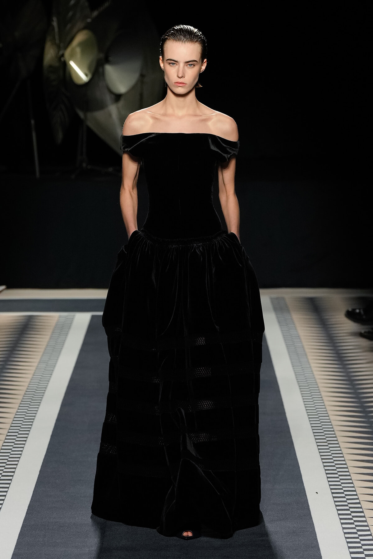 Model on a runway wearing a black off‑the‑shoulder velvet gown with a fitted bodice and full floor‑length skirt.