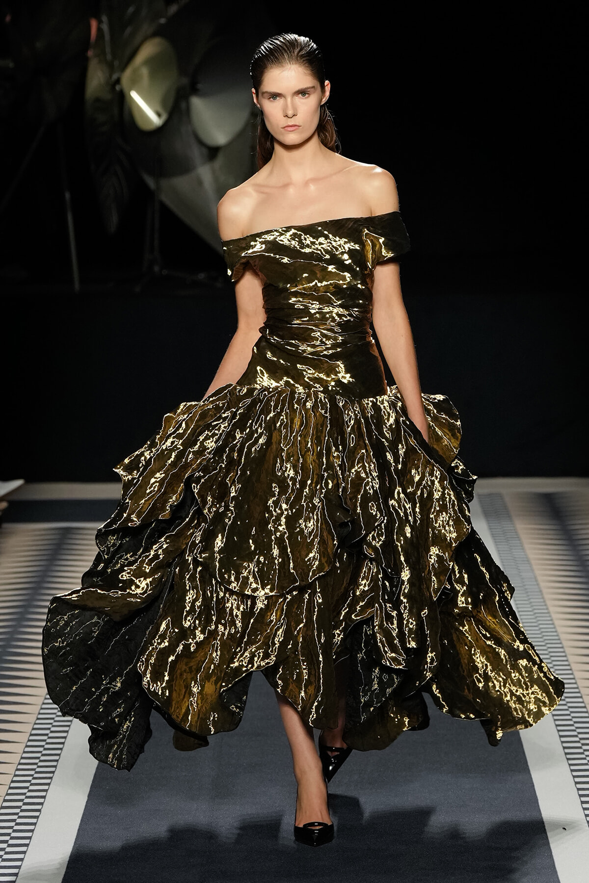Model wearing an off-the-shoulder gold and black metallic ballgown walking the runway.