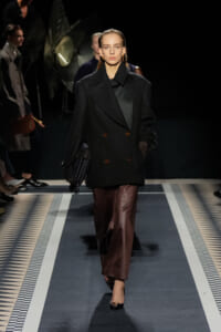 Model walks a runway in a black oversized double-breasted coat, brown satin skirt, and fringe handbag under spotlights.
