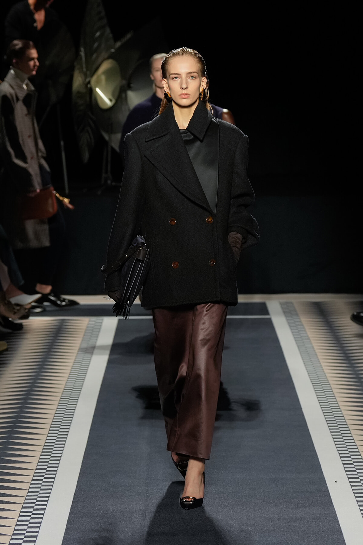 Model walks a runway in a black oversized double-breasted coat, brown satin skirt, and fringe handbag under spotlights.