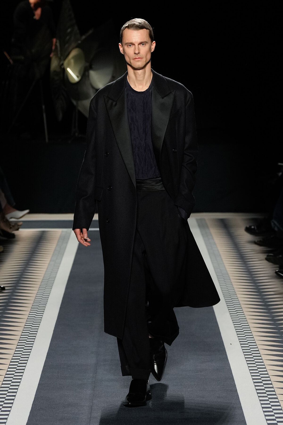 Male model walking a fashion runway in a long black overcoat over a dark sweater and wide-leg trousers on a patterned catwalk.