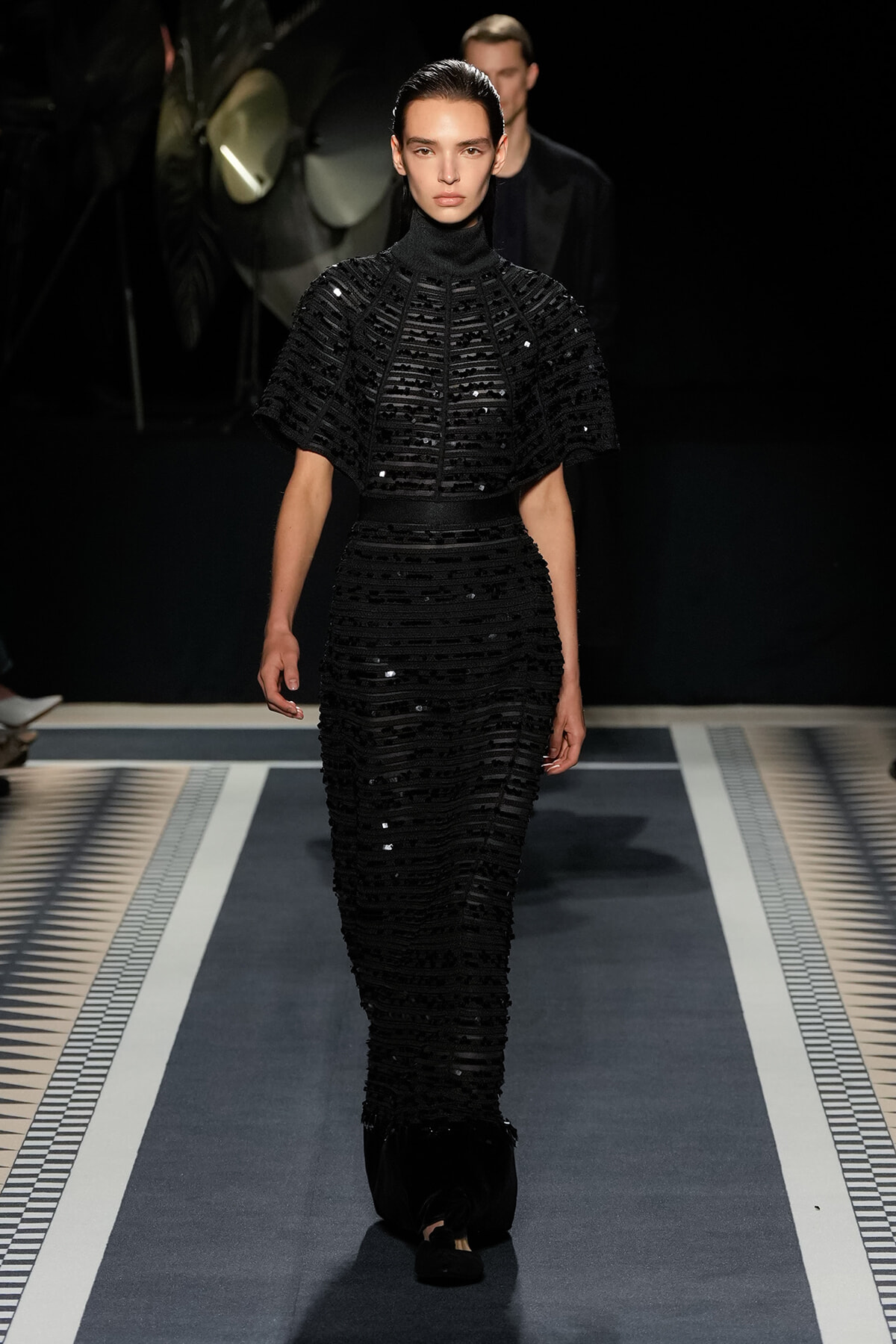 Model in a black sequined high-neck cape and matching beltless gown walking the runway.