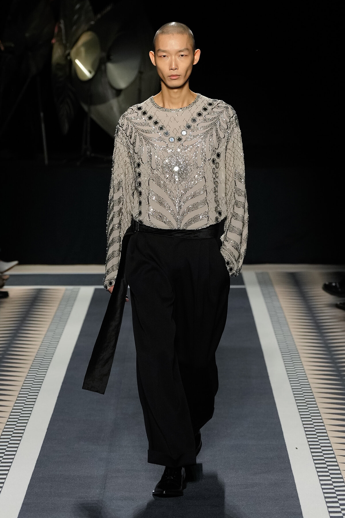Male model walks a fashion-show runway in a beige sheer top adorned with silver beadwork and a long black sash, paired with black wide-leg trousers.