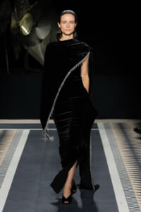 Model walks a fashion runway in a black velvet draped gown with a silver-fringed diagonal cape, exposing one leg and wearing open-toe black heels.