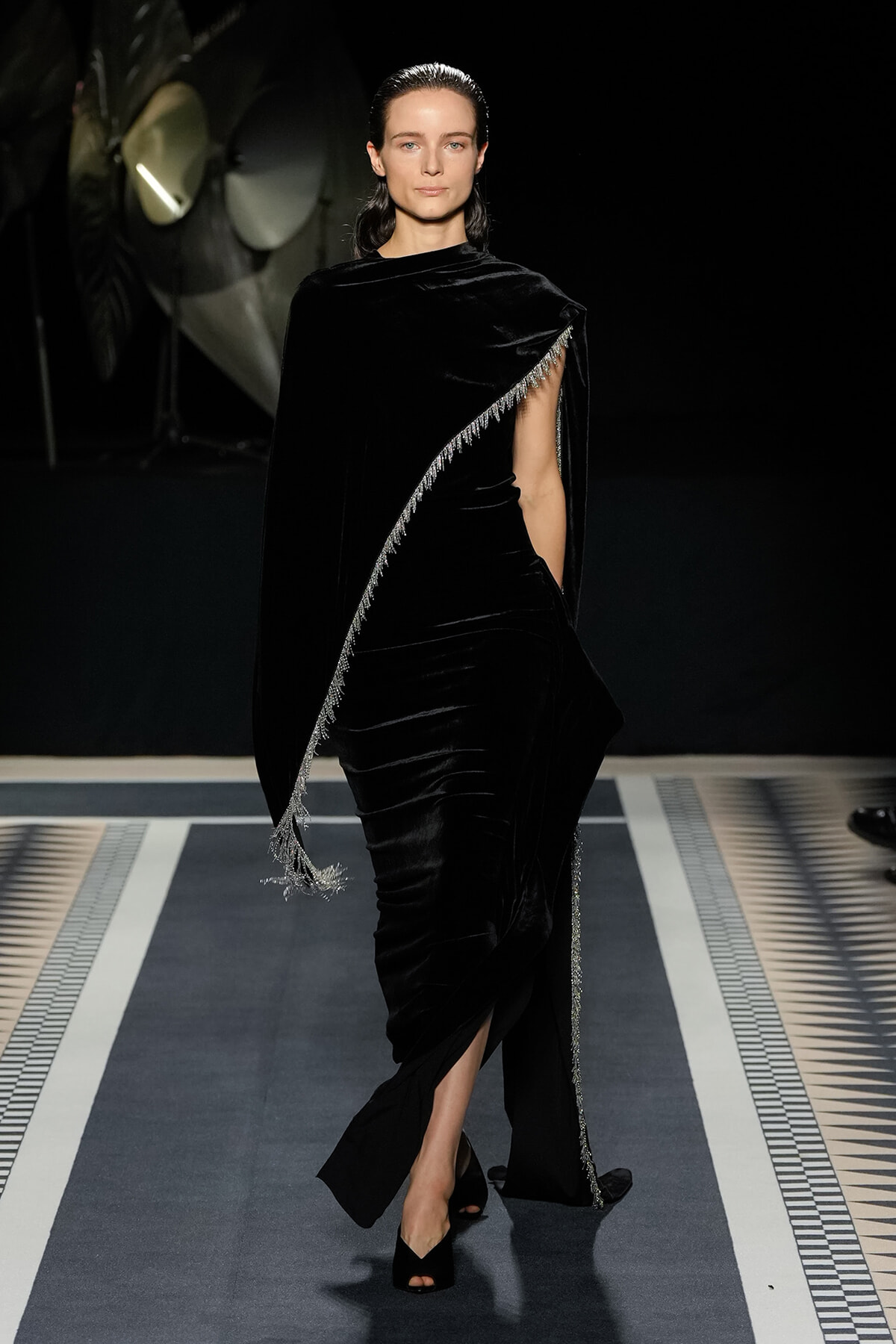 Model walks a fashion runway in a black velvet draped gown with a silver-fringed diagonal cape, exposing one leg and wearing open-toe black heels.