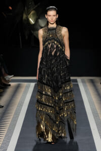 Model walks the runway in a black-and-gold lace gown with layered, metallic detailing and sheer panels at the top.