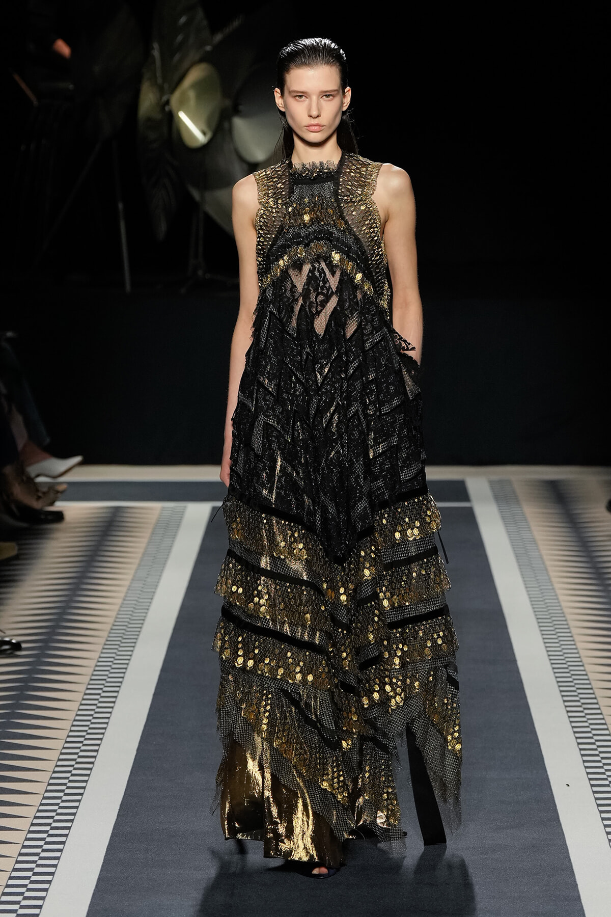 Model walks the runway in a black-and-gold lace gown with layered, metallic detailing and sheer panels at the top.