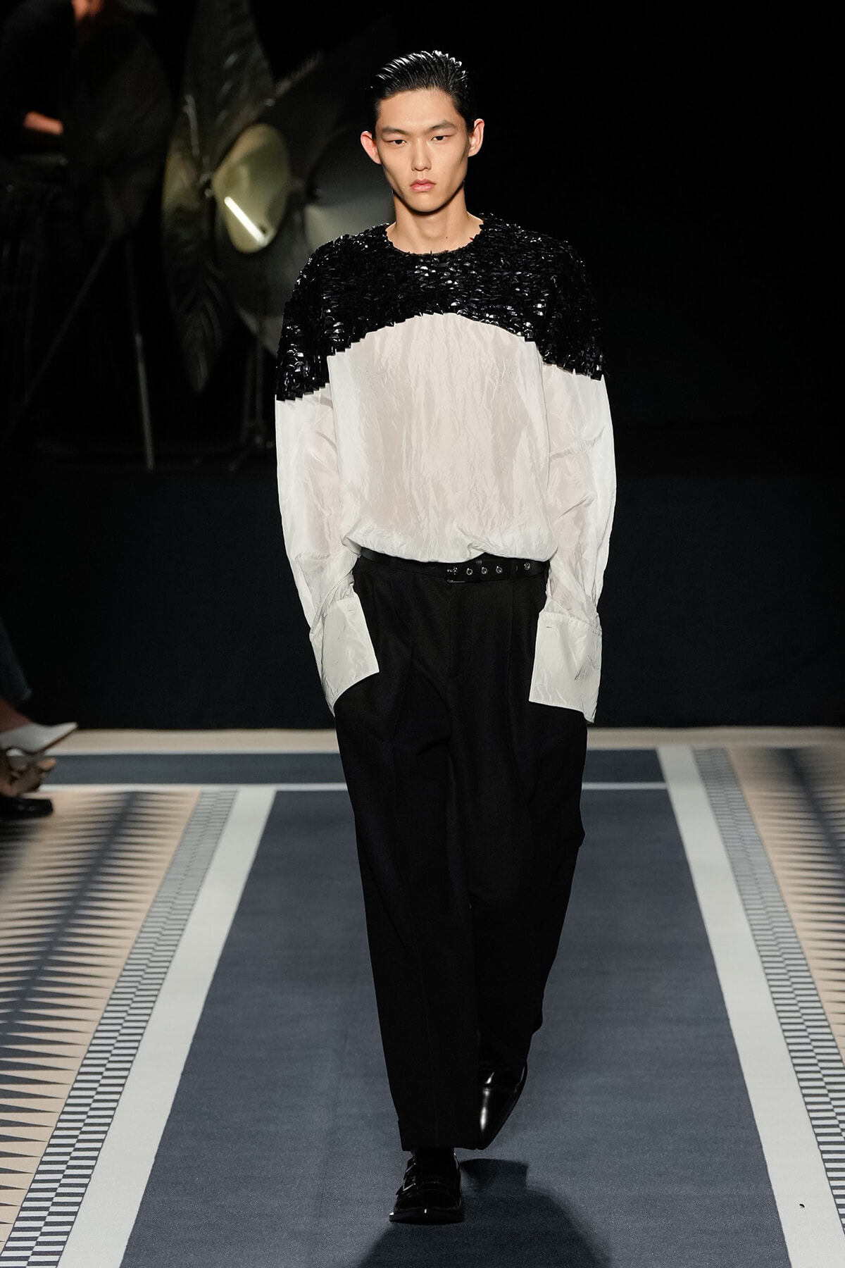 Male model walks a runway in a two-tone black-and-white top with a sequined black upper panel, white crinkled lower section, black trousers, and glossy shoes.