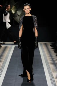 Runway model in a floor-length black velvet gown with sculpted metallic cape sleeves and black gloves, walking the catwalk.