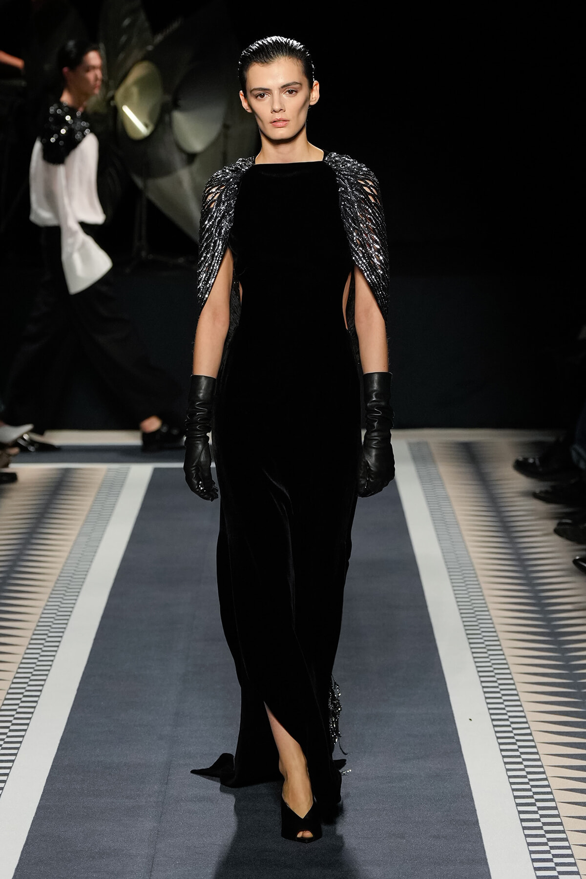 Runway model in a floor-length black velvet gown with sculpted metallic cape sleeves and black gloves, walking the catwalk.