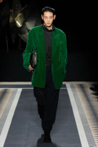 Male model walking a runway in a vivid green velvet blazer over black outfit, clutch in hand.