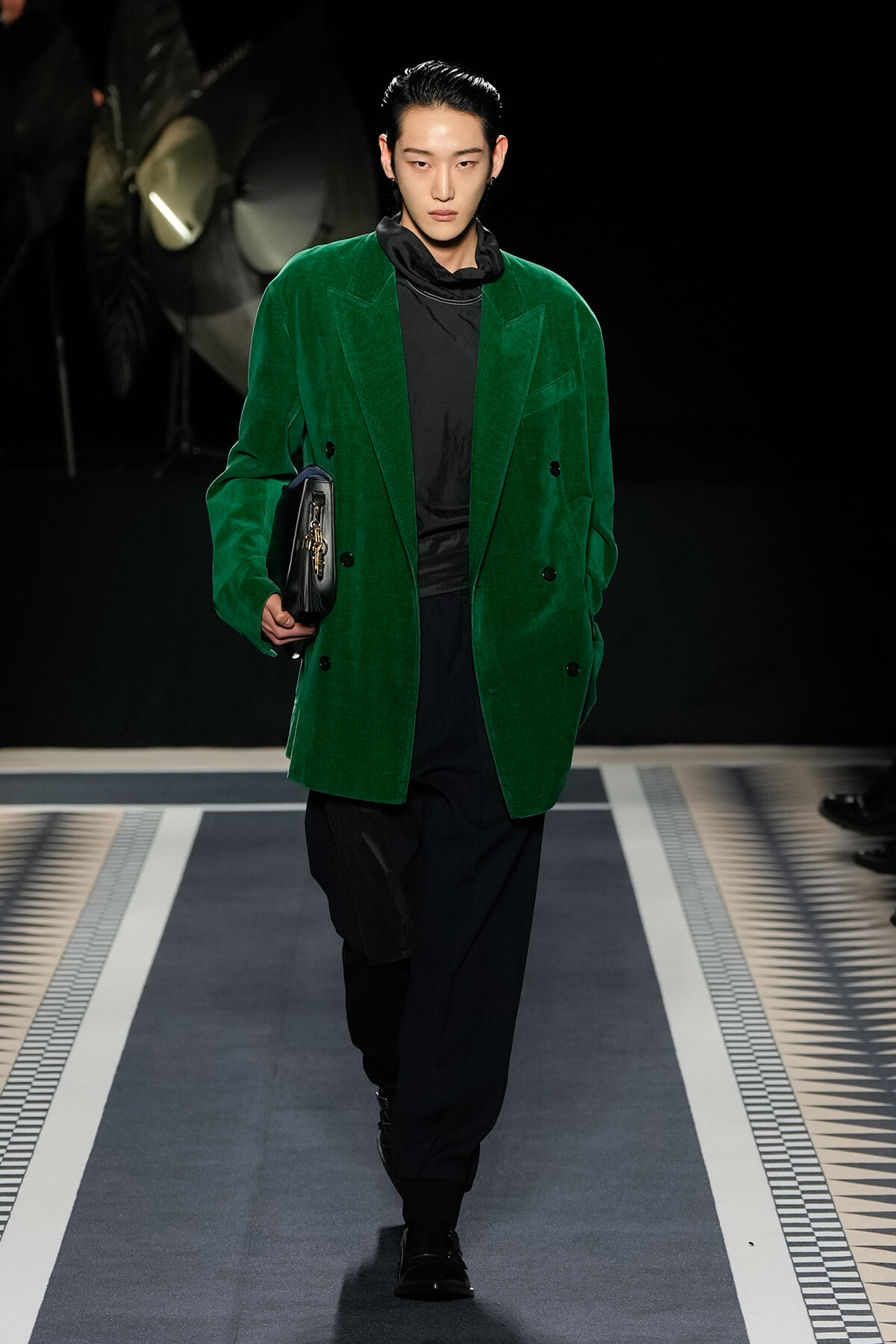 Male model walking a runway in a vivid green velvet blazer over black outfit, clutch in hand.