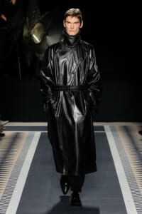 Model walking the runway in a black leather trench coat with a high collar and belted waist, hands in pockets, low-contrast background.