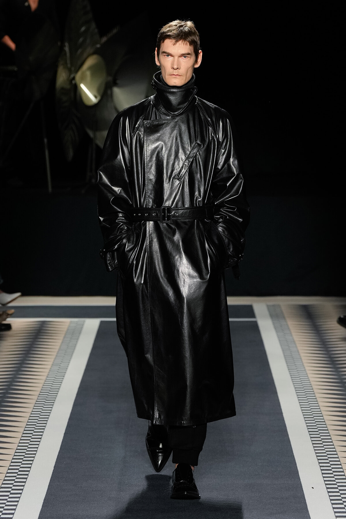 Model walking the runway in a black leather trench coat with a high collar and belted waist, hands in pockets, low-contrast background.