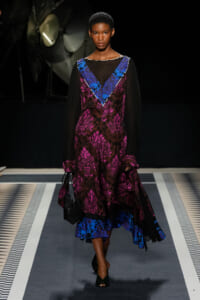 Model walking a runway in a sheer black-sleeve dress with magenta floral print and blue V-neck trim, carrying a small black bag.