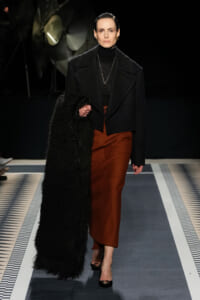 Model on a runway wearing a black turtleneck and oversized black coat with a rust-colored midi skirt, grasping a long black textured scarf.