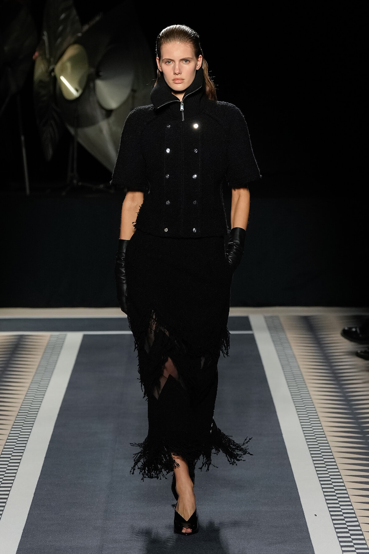 Model walking the runway in a black textured two‑piece ensemble: cropped double‑breasted jacket and fringed skirt, with black gloves.