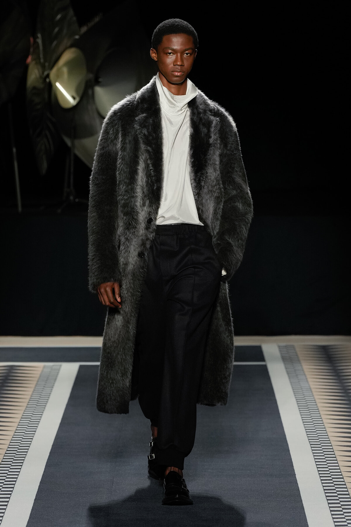 Male model on a runway wearing a long dark fur coat over a white turtleneck and black trousers.
