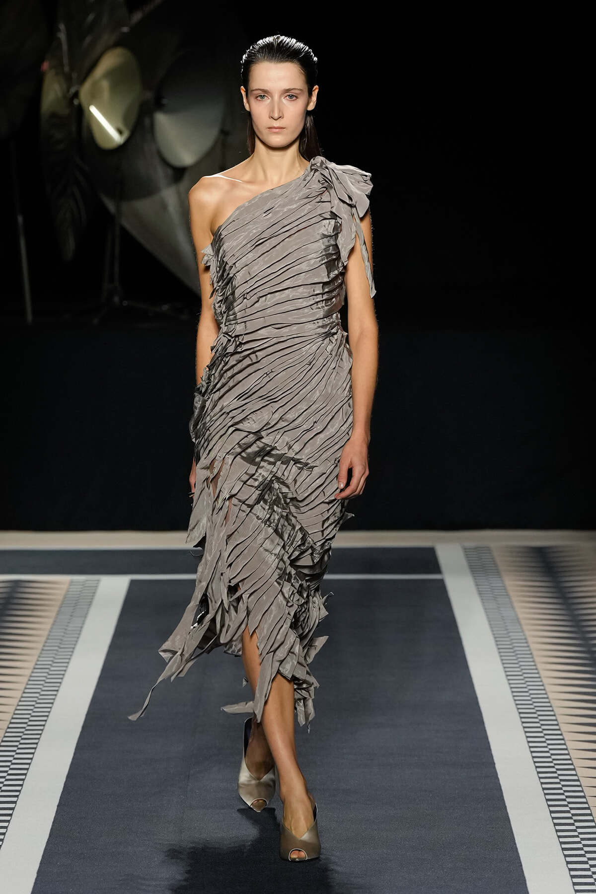 Model walking a runway in a one-shoulder gray, pleated dress with layered ruffles