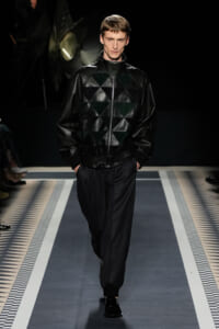 Male model walking a runway in a black quilted leather jacket and black trousers at a fashion show.
