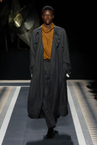 Male model on a runway wearing a long charcoal trench over a rust-brown top, black pants, and black shoes against a dark backdrop.