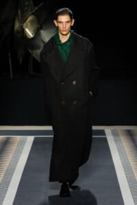Male runway model wearing a long black double-breasted coat with brown buttons and a green top, walking the catwalk.