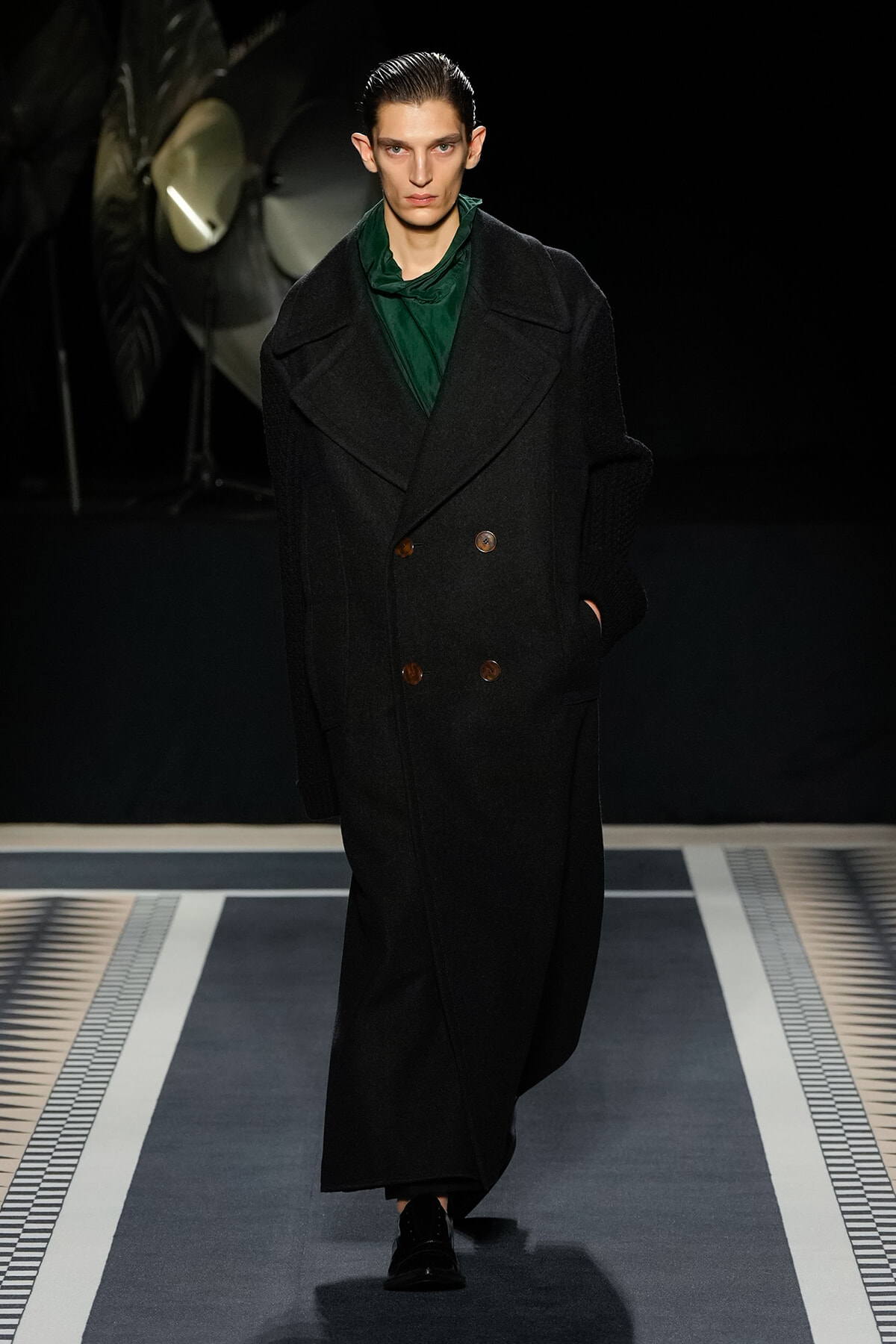 Male runway model wearing a long black double-breasted coat with brown buttons and a green top, walking the catwalk.