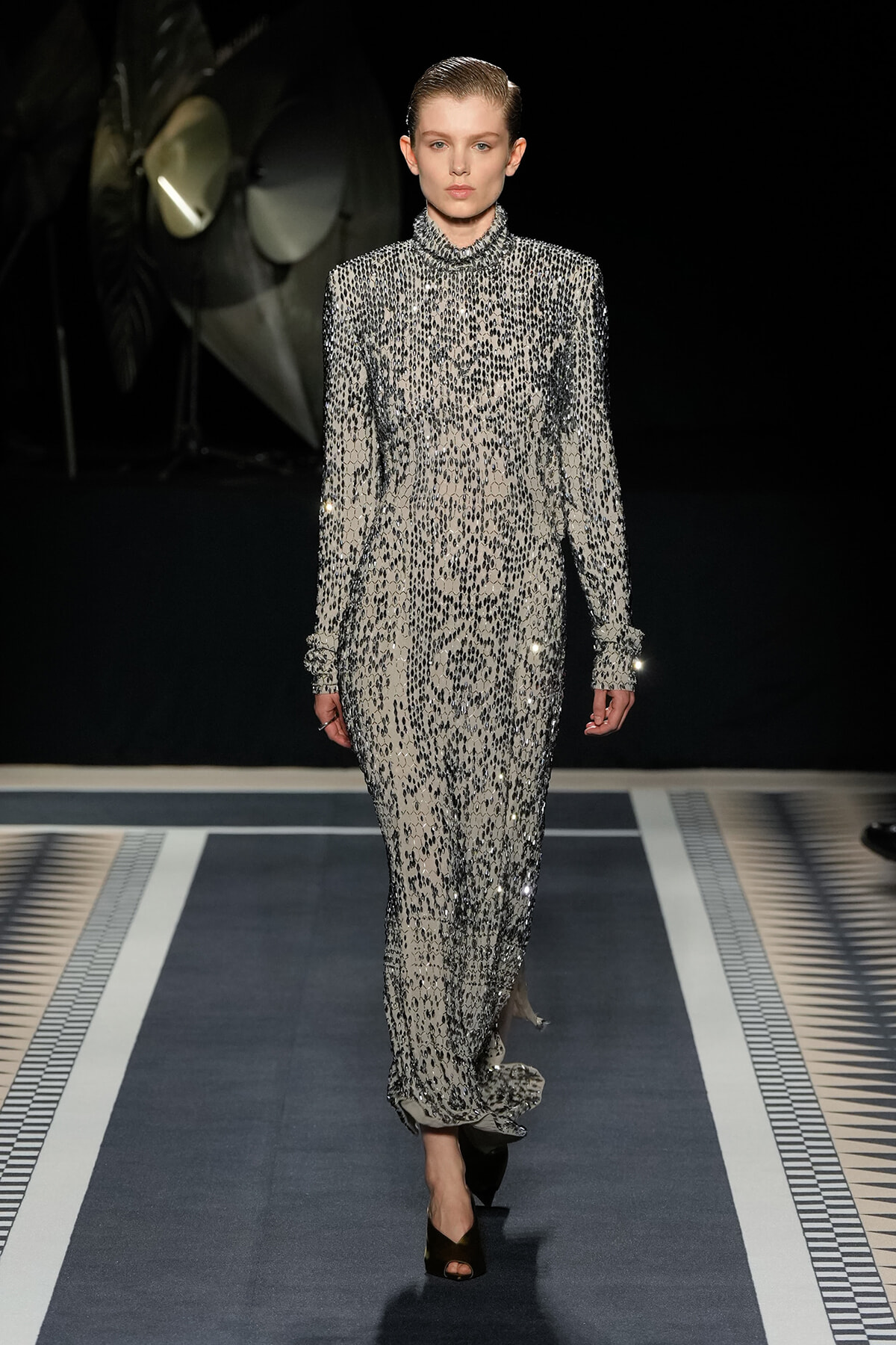 Model walking the runway in a floor-length silver and black beaded gown with a high neckline and long sleeves, on a dark fashion show set.