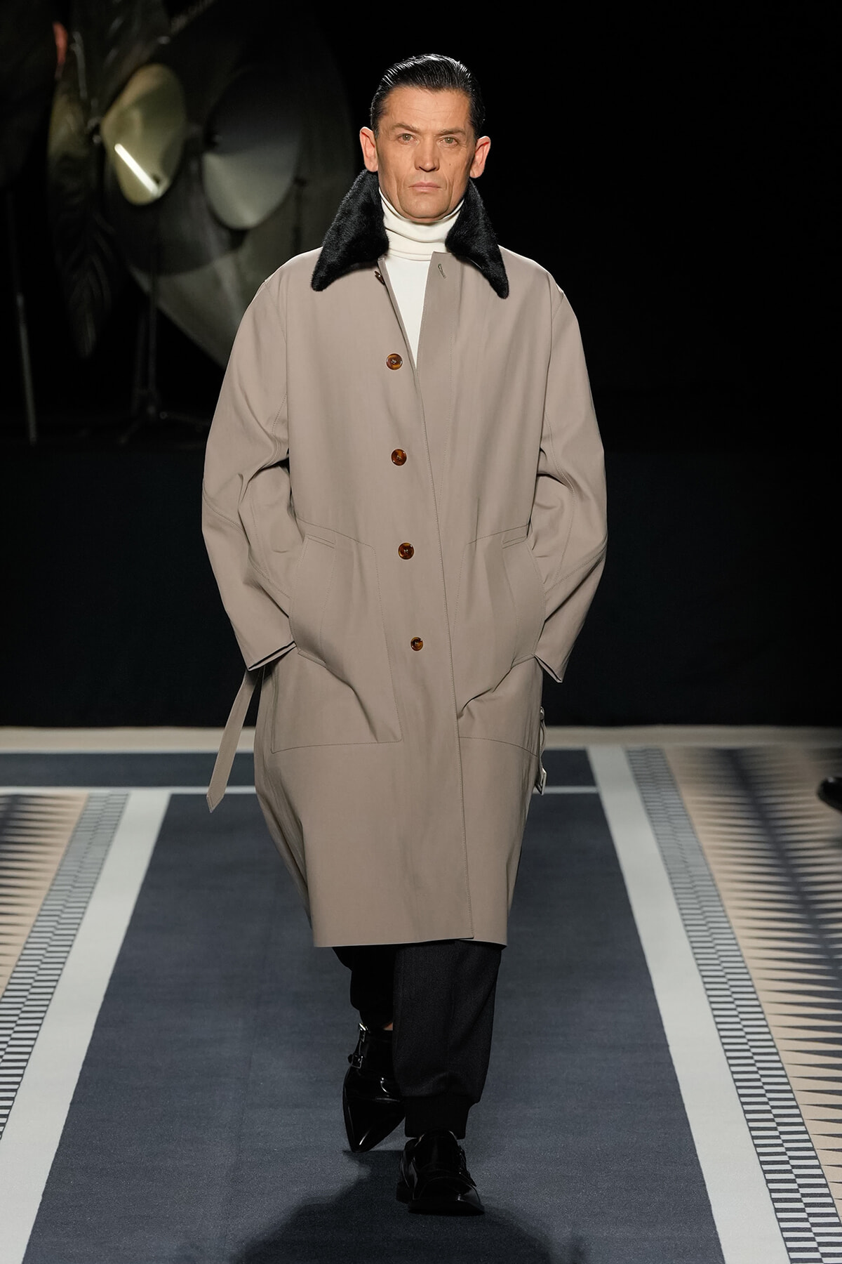 Male model on a runway wearing a beige trench coat with a black fur collar, white turtleneck, and black trousers with glossy shoes.