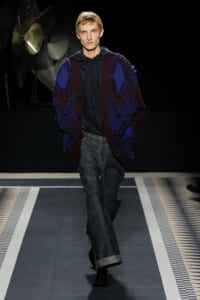 Male model walking runway in oversized maroon and blue cardigan with bold chevron knit, navy hoodie, wide dark denim jeans, black shoes